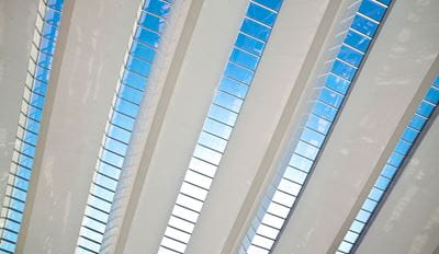 Modern building interior with white structural beams and diagonal glass skylight panels revealing a blue sky