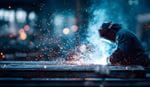 A welder in protective gear crouches over metal plates, with bright sparks and blue smoke filling a dark industrial workshop