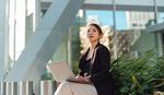 A woman in business attire sits outdoors with a laptop, surrounded by plants, beneath modern architecture. 