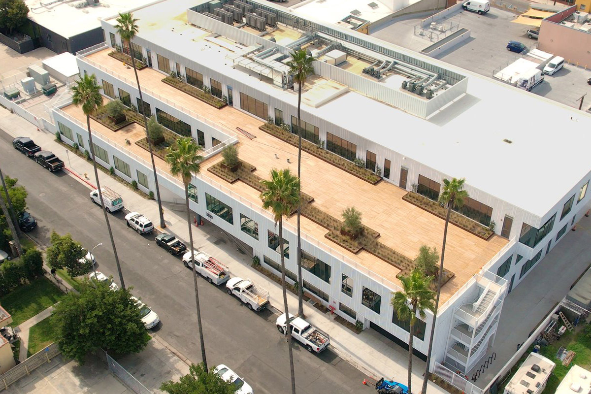 Modern white building with wooden rooftop terrace and palm trees, viewed from above.